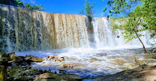 A cascading waterfall flows over a rocky ledge, surrounded by lush greenery and a clear blue sky.
