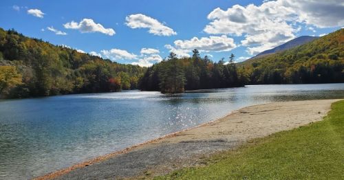 A serene lake surrounded by trees and mountains, with a sandy shore and a clear blue sky dotted with clouds.