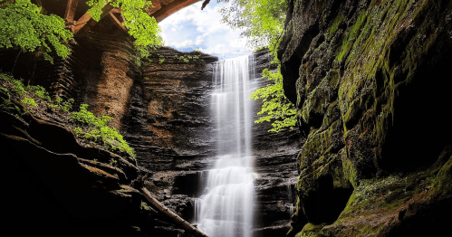 A serene waterfall cascades down rocky cliffs, surrounded by lush greenery and dappled sunlight.