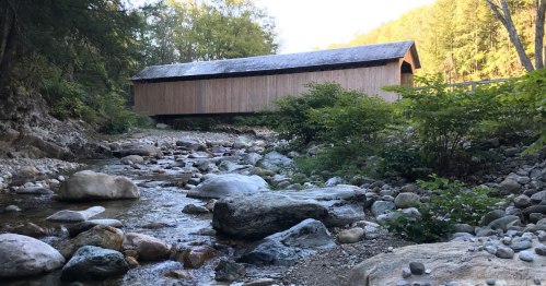 A wooden covered bridge spans a rocky stream, surrounded by trees and hills in a serene natural setting.