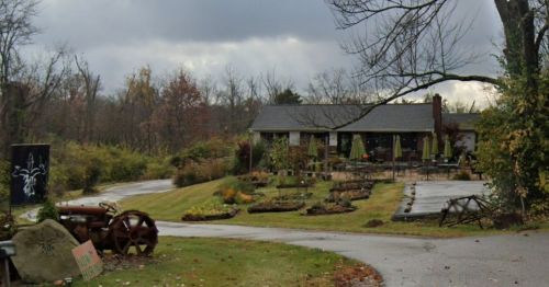 A rustic building surrounded by gardens and trees, with a cloudy sky overhead and a winding driveway.