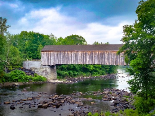 A wooden covered bridge spans a river, surrounded by lush greenery and a cloudy sky. Rocks are visible in the water.