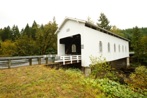 A white covered bridge stands over a small stream, surrounded by trees and greenery on a cloudy day.