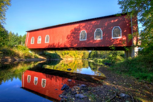 A red covered bridge spans a calm river, reflecting in the water, surrounded by lush greenery and a clear blue sky.