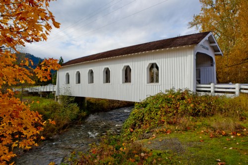 A white covered bridge spans a stream, surrounded by vibrant autumn foliage and a cloudy sky.