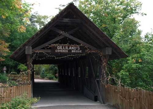 A wooden covered bridge with a sign reading "Gilliland's Covered Bridge," surrounded by greenery and fairy lights.