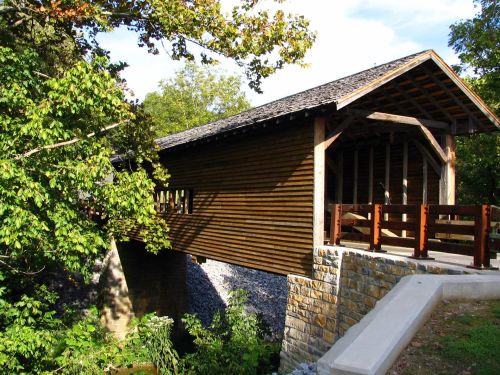 A wooden covered bridge surrounded by trees, with a stone foundation and a clear blue sky in the background.