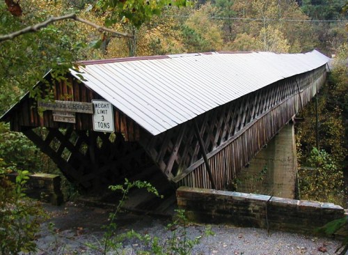 A wooden covered bridge with a peaked roof spans a small creek, surrounded by trees and autumn foliage.