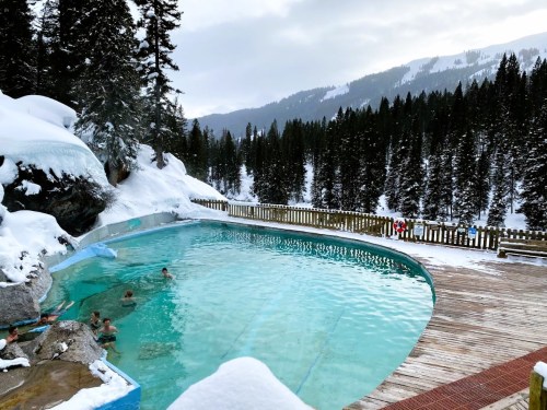 A scenic outdoor hot spring pool surrounded by snow-covered trees and mountains, with people enjoying the water.