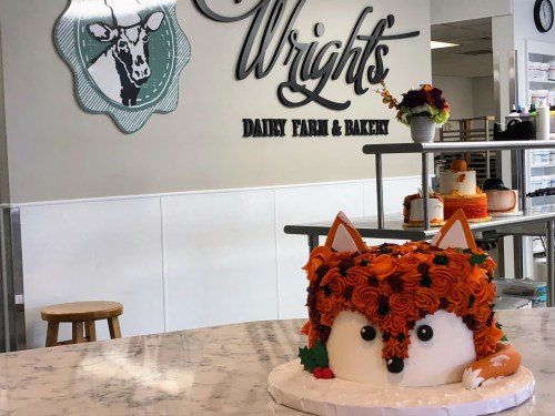 A whimsical fox-shaped cake with orange frosting sits on a marble counter in a bakery, with a dairy farm sign in the background.