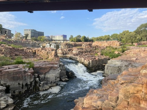 A scenic view of a waterfall cascading over rocky terrain, with buildings and trees in the background under a blue sky.