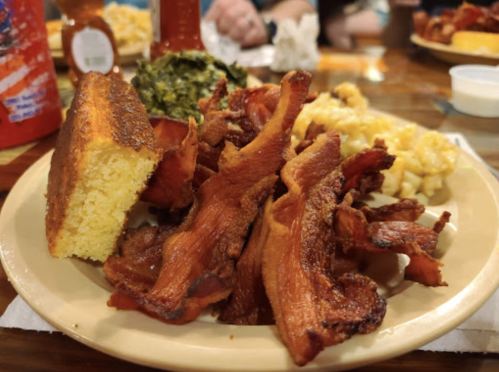 A plate of crispy bacon, cornbread, collard greens, and macaroni and cheese on a wooden table.