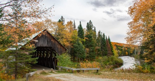 A rustic covered bridge surrounded by vibrant autumn foliage and a winding river under a cloudy sky.