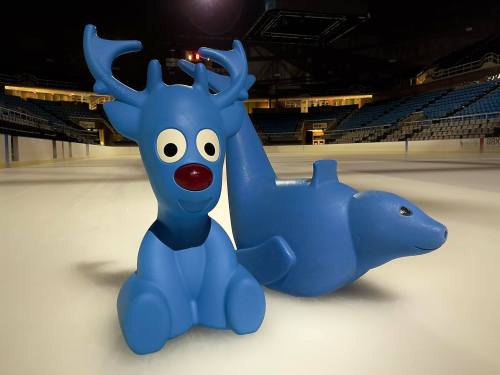 Two colorful plastic toys on an ice rink: a blue reindeer and a blue seal, set against empty bleachers.