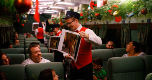 A train conductor in a festive outfit shows photos to passengers in a decorated train car filled with holiday decor.