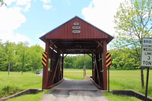 A red covered bridge with a sign reading "The Kiepps Bridge," surrounded by green fields and trees under a blue sky.