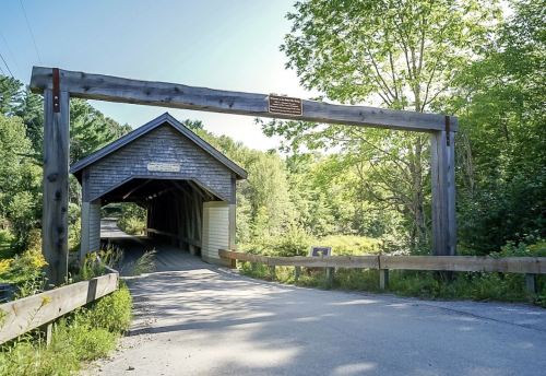 A scenic view of a covered bridge with a wooden entrance, surrounded by lush greenery and a clear blue sky.