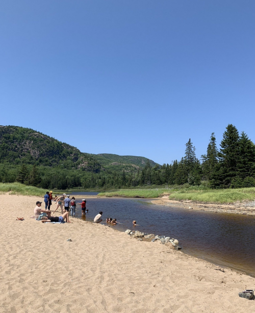 A sandy riverbank with people enjoying the outdoors, surrounded by green hills and clear blue skies.