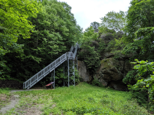 A metal staircase leads up a rocky hillside, surrounded by lush green trees and grass.