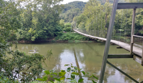 A serene river scene with a suspension bridge and a small boat on the water, surrounded by lush greenery.