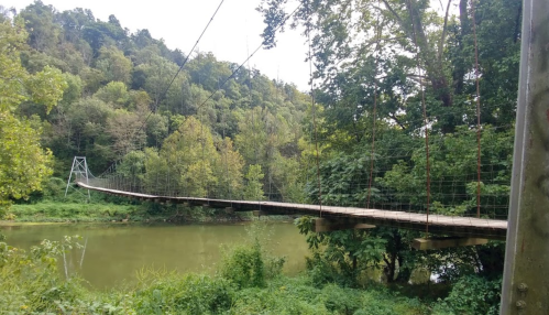 A wooden suspension bridge spans a calm river, surrounded by lush green trees and hills.