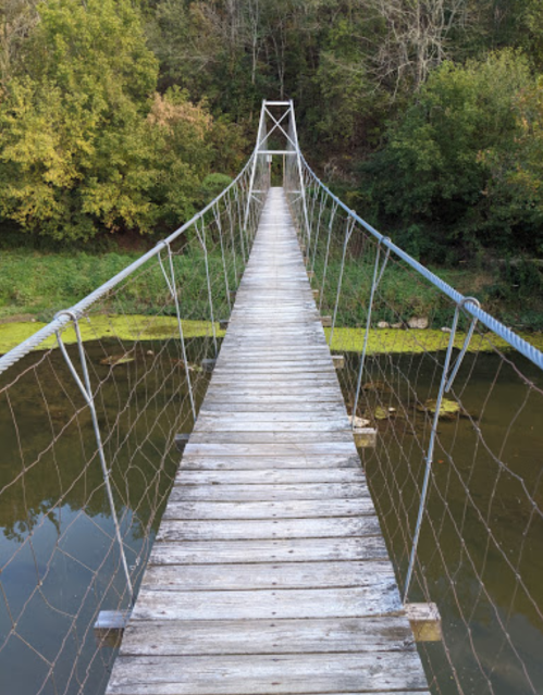 A wooden suspension bridge stretches over a calm river, surrounded by lush green trees.
