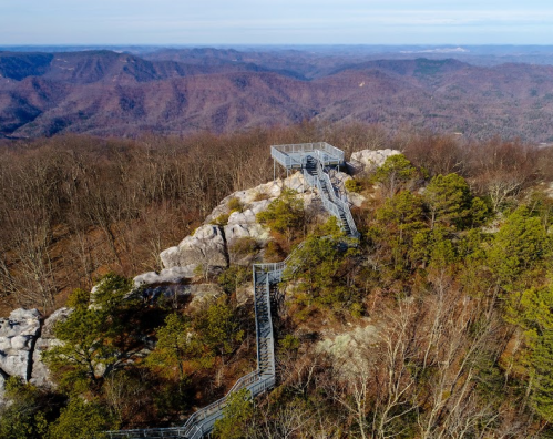 A scenic overlook with a metal walkway on a rocky outcrop, surrounded by trees and mountains in the background.