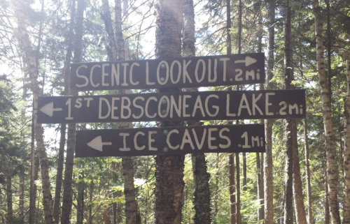 Wooden signpost in a forest with directions to Scenic Lookout, Debsconeag Lake, and Ice Caves, with distances marked.