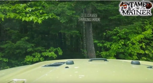 View from a vehicle approaching a sign for the Ice Caves parking area, surrounded by lush green trees.