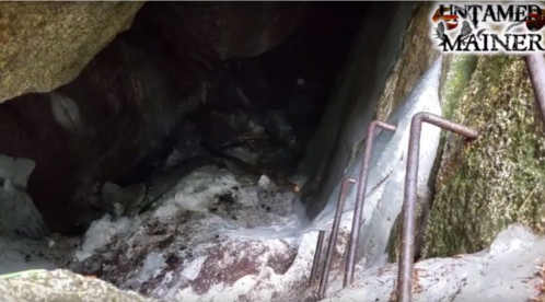 A dark cave entrance with icy steps leading down, surrounded by large rocks and snow.