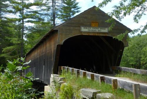 A wooden covered bridge surrounded by trees, with a sign reading "Hemlock Covered Bridge, Fryeburg 1857."