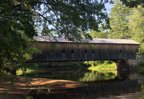 A wooden covered bridge spans a calm river, surrounded by lush greenery and trees under a clear blue sky.