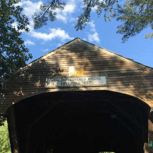 A wooden covered bridge with a sign reading "Hemlock Covered Bridge Fryeburg 1857" under a blue sky and trees.