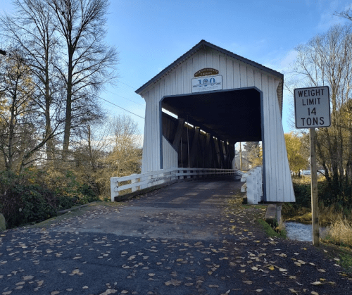 A white covered bridge with a sign indicating a 14-ton weight limit, surrounded by trees and fallen leaves.
