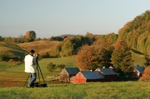 A photographer captures a scenic landscape of rolling hills and colorful trees near a rustic farm.