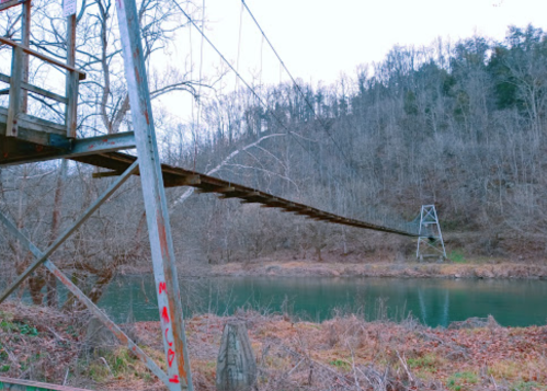 A suspension bridge stretches over a calm river, surrounded by bare trees and a hilly landscape in the background.