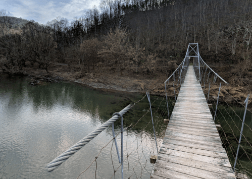 A wooden suspension bridge stretches over a calm river, surrounded by bare trees and a hilly landscape.