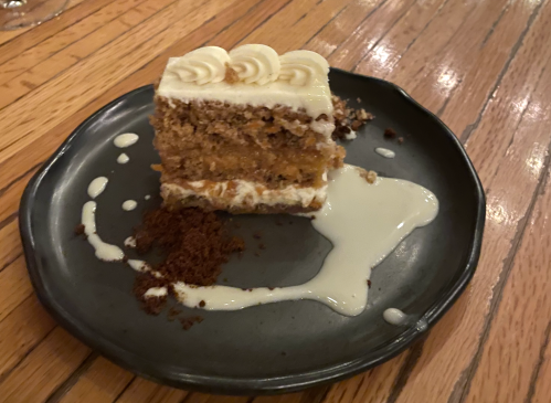 A slice of layered cake with cream frosting on a dark plate, accompanied by a drizzle of sauce and crumbs.