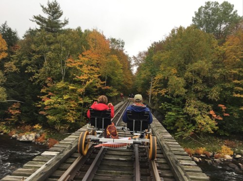 Two people ride a rail bike on a wooden train track surrounded by colorful autumn trees.