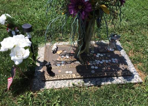 Gravestone of John Paycheck, adorned with flowers, coins, and decorative items, set in a grassy area.