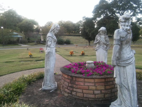 Three weathered statues stand around a flower-filled stone planter in a serene cemetery setting.
