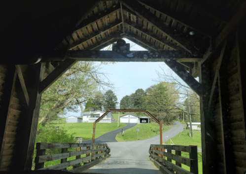 View from inside a covered bridge, leading to a winding road and houses in the distance, surrounded by trees.