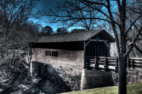 A wooden covered bridge spans a rocky creek, surrounded by bare trees and a clear blue sky.