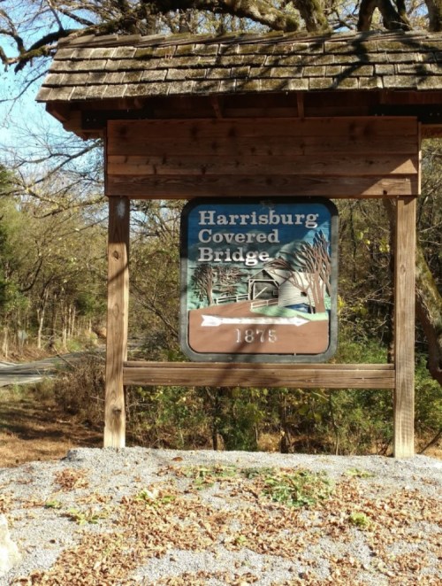 Sign for Harrisburg Covered Bridge, featuring a painted image of the bridge and the year 1875. Surrounded by trees.