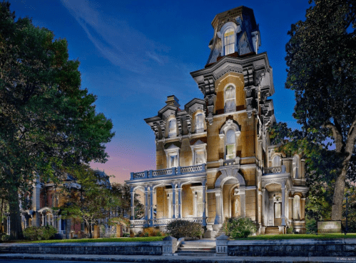 A grand, historic mansion with intricate architecture, illuminated at dusk, surrounded by lush trees and gardens.