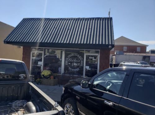 A small storefront with a black roof, featuring a sign for "Sheila's Place" and parked vehicles in front.