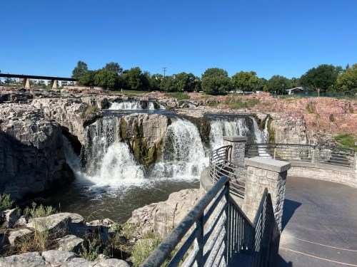 A scenic view of a waterfall cascading over rocky cliffs, with a clear blue sky and greenery in the background.