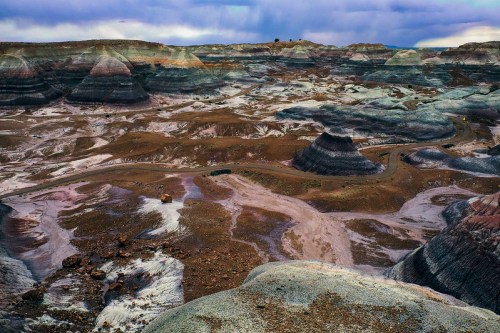 A vast, colorful landscape of eroded hills and valleys under a cloudy sky, showcasing unique geological formations.