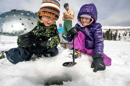 Two children in winter gear are ice fishing, focused on a hole in the snow, with adults in the background.