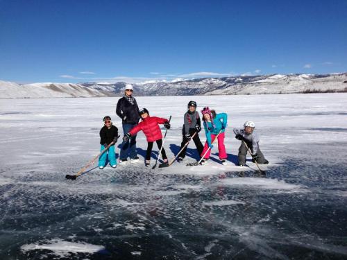 A group of children and an adult play hockey on a frozen lake, surrounded by snow-covered mountains under a clear blue sky.